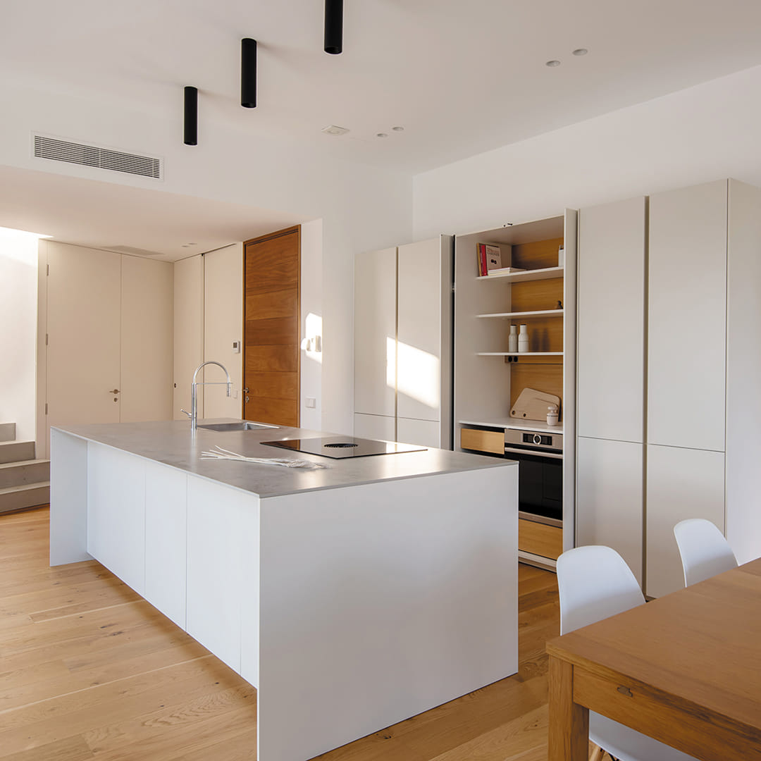 Cocina blanca integrada en zona de día con armarios en columna con puertas escamoteables.