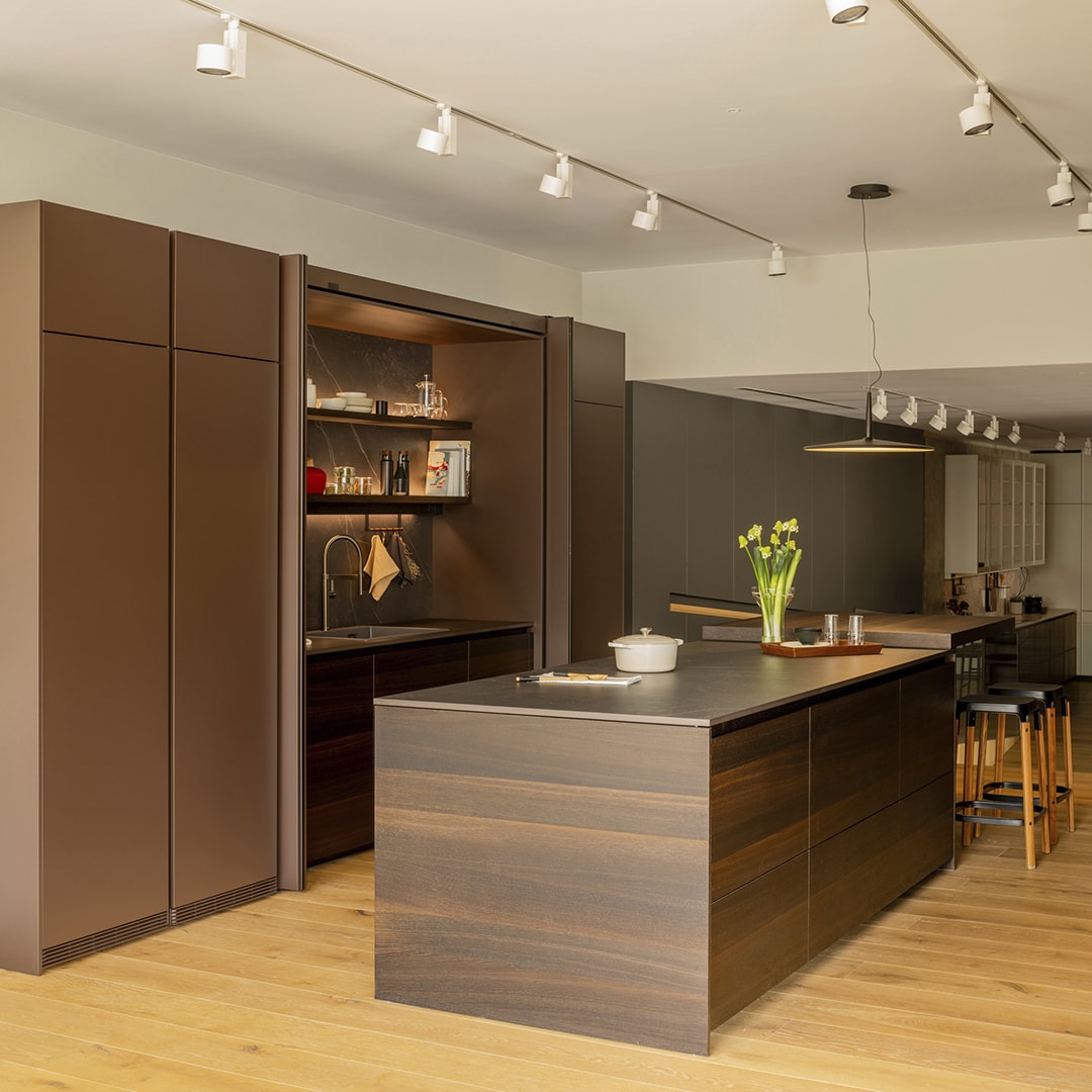 Cocina en color marrón oscuro con acabados en madera en la exposición de Santos en Santiago Interiores.